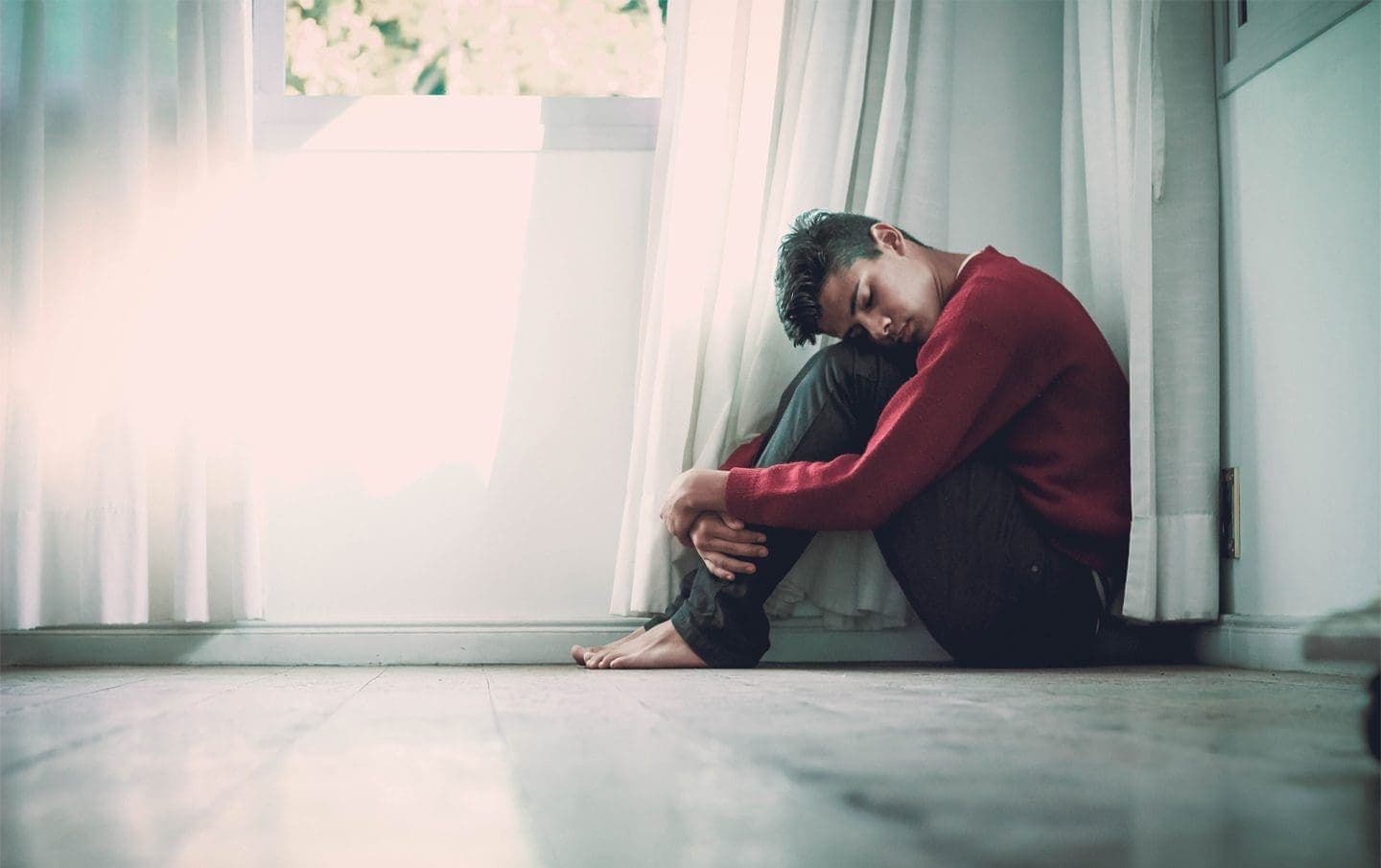 young man sitting while leaning his head on his knees below the window